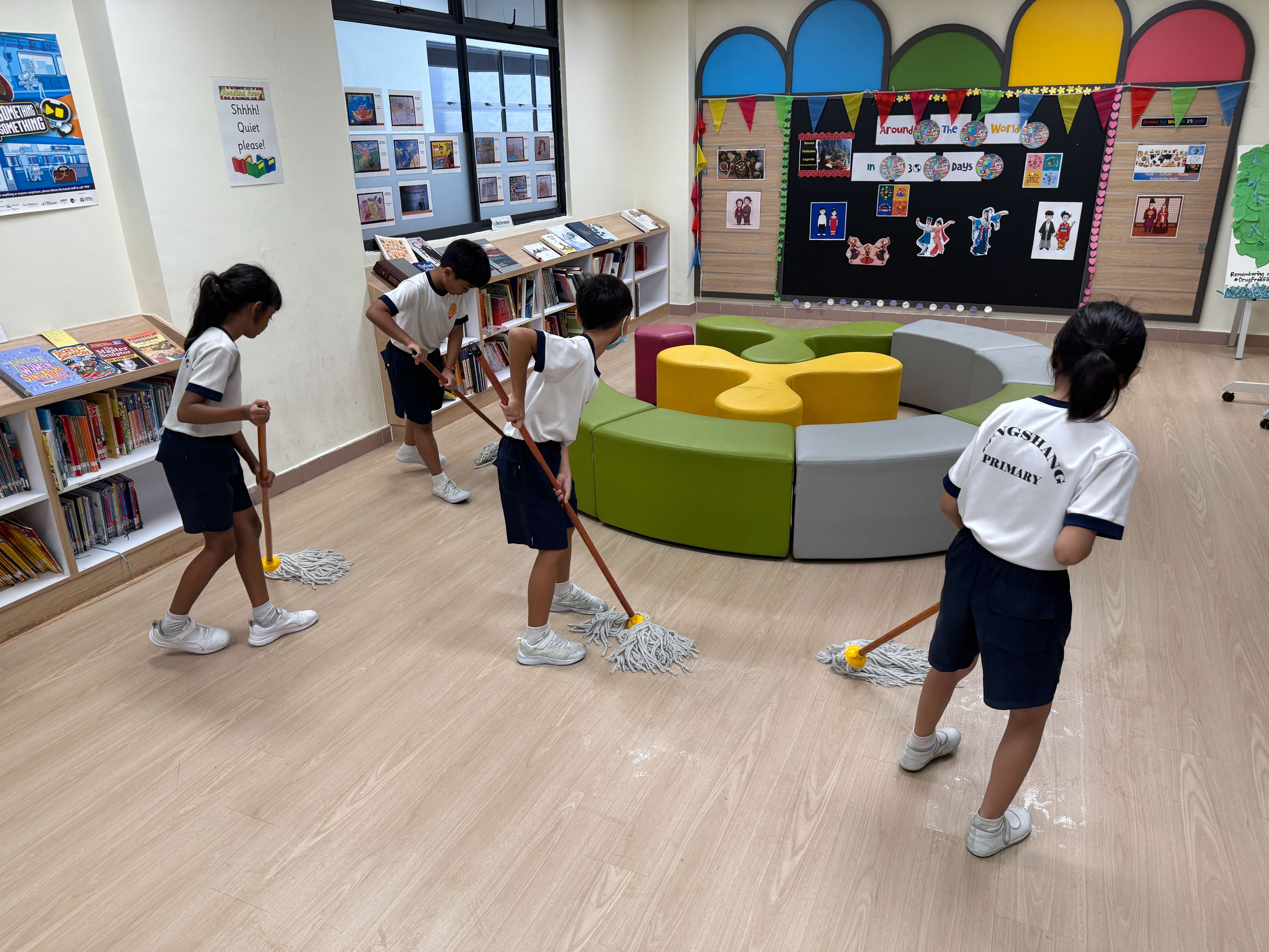 Four Gongshang Primary School students in white and navy blue uniforms mopping the floor of a school library. Bookshelves lined with books are visible along the walls, alongside a colourful circular seating arrangement and a decorated display board in the background.